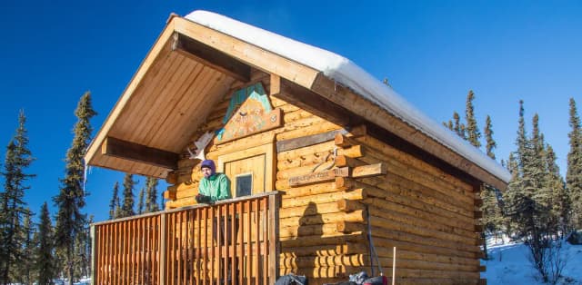 A woman looks out from the front deck of a log cabin.