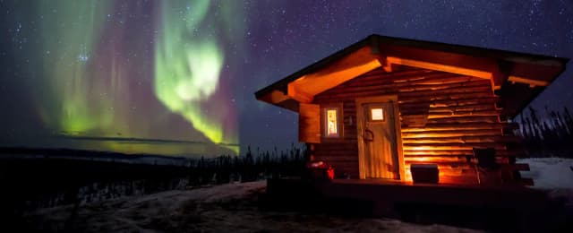 A log cabin stands on a hill in front of northern lights on a starry sky.