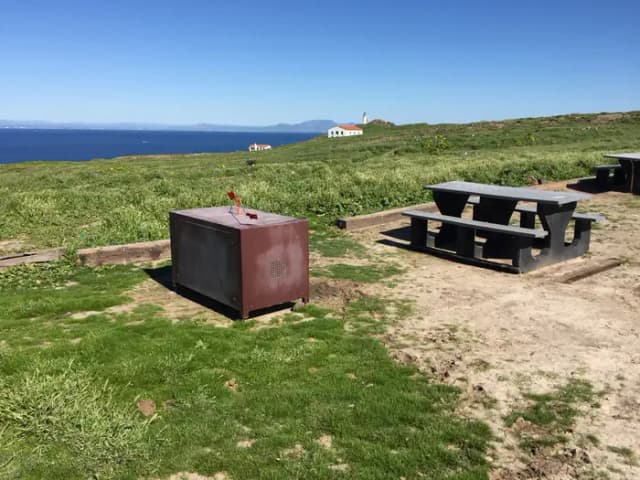 Picnic table and metal food storage box on terrace overlooking ocean, two buildings and lighthouse.