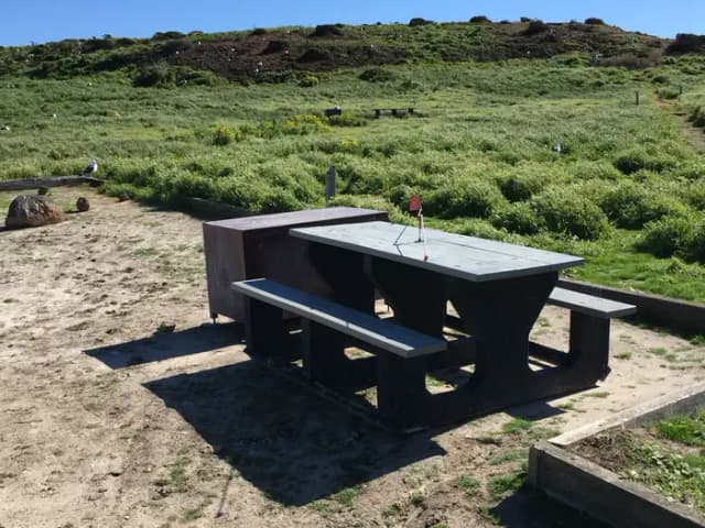 Picnic table and metal food storage box on terrace overlooking ocean, two buildings and lighthouse.
