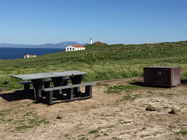 Picnic table and metal food storage box on terrace overlooking ocean, two buildings and lighthouse.