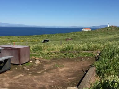 Picnic table and metal food storage box on terrace overlooking ocean, two buildings and lighthouse.