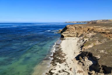 Sandy beach with ocean and small waves and steep coastal bluffs covered in dry grass., filed by the agency against 10 campsites at Santa Rosa Island Backcountry Beach Camping