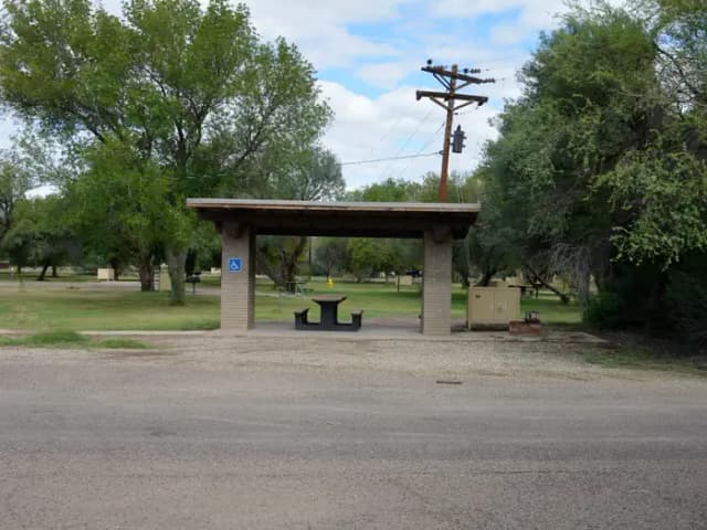Wide view of the shade shelter, picnic table, and bear box for Site #14. The shelter, picnic table, and bear box are built on a paved area to provide ease of access to the site's amenities. The gravel driveway is directly in front of the shade shelter, allowing the shortest distance possible between a parked vehicle and the campsite. A grassy field with cottonwood trees and a telephone pole can be seen just behind the site.
