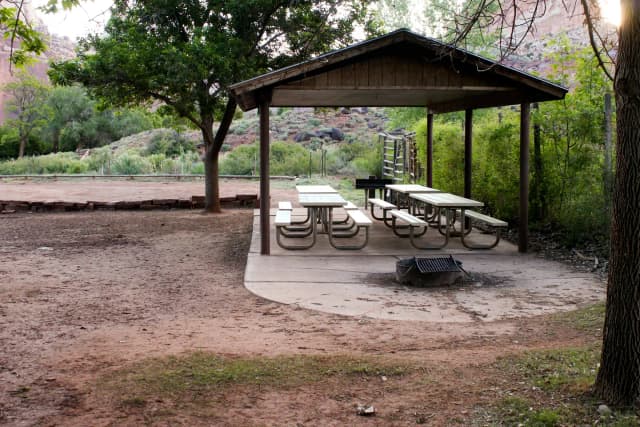 A pavilion covers four white picnic tables on pavement. There is a fire pit to the right of the pavilion, still on pavement. The pavilion backs up to foliage. A patch of dirt is in front of it and the fire pit