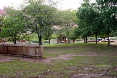 A grassy field. A fence is in the front of image, across half of the image. There are a few trees in the area. A small building is in the background.