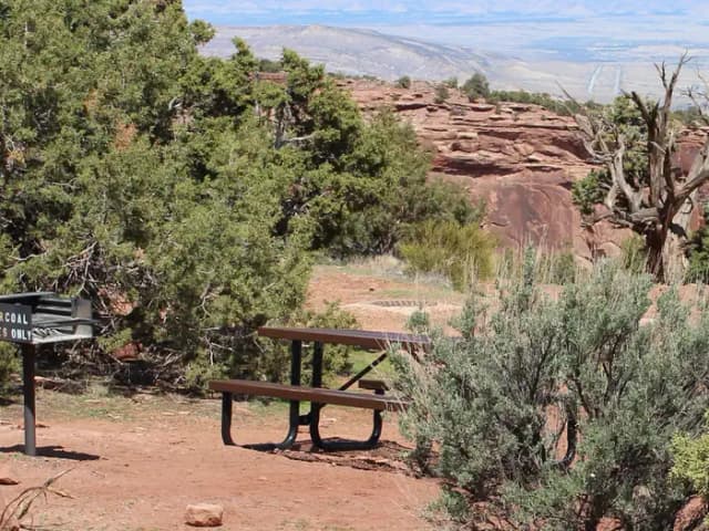 Picnic table and grill at Loop B Site 37 near a red rock canyon edge, surrounded by juniper trees and desert brush with expansive views
