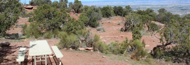A campsite with a light-colored picnic table on a red dirt clearing, surrounded by desert shrubs and trees. The site overlooks a wide valley in the background with distant mountain ridges under a clear sky.