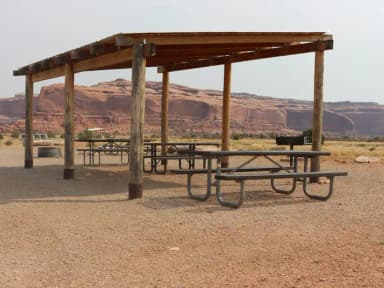Lone Mesa Group Site E shade shelter, picnic tables, and fire ring. Tall, red rock plateaus line the horizon.