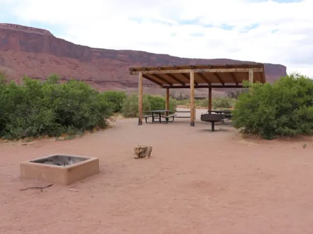A close up of the Lower Onion Creek Group Site A shade shelter, picnic tables, standing grill, and hang out area next to fire pit. Vegetation lines the area and in the distance are towering, red rock cliffs.