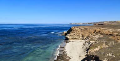 Sandy beach with ocean and small waves and steep coastal bluffs covered in dry grass., filed by the agency against 2 campsites at Santa Rosa Island Backcountry Beach Camping