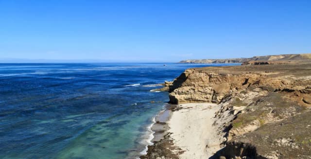Sandy beach with ocean and small waves and steep coastal bluffs covered in dry grass.