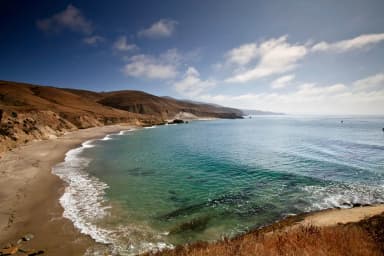 Sandy beach with ocean and small waves and steep coastal bluffs covered in dry grass., filed by the agency against 3 campsites at Santa Rosa Island Backcountry Beach Camping