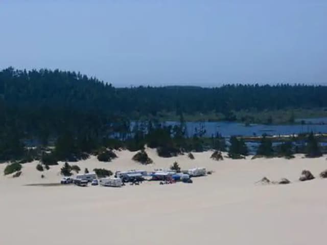 Group of trailers and trucks on a large expanse of sand near a conifer encircled lake under a blue sky.