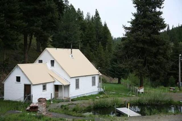 White house on flat green grass between a conifer covered hill and a small pond with a dock.