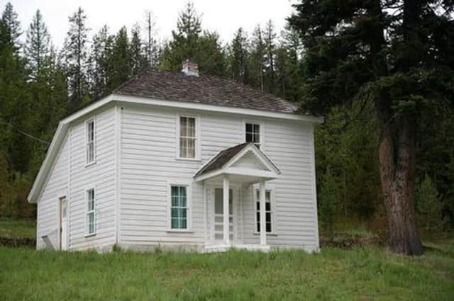White, two-story house on a grassy hill next to a fir forest.